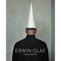 A photograph showing the back of a person with short hair, wearing a black jacket and a tall, white, cone-shaped paper hat. The person stands against a textured gray wall. White text on their back reads ERWIN OLAF and STRANGE BEAUTY.
