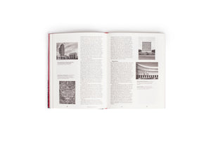 An open book with a red binding, seen from above against a white background. The pages contain German text and are illustrated with black-and-white photographs of 20th-century architecture, including buildings and an aerial city plan.