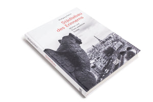 A white hardcover book, Städtebau des Erinnerns by Wolfgang Pehnt, lies on a white surface. The cover features a black and white photo of a stone gargoyle looking out over a cityscape with the Eiffel Tower. The title is printed in red.