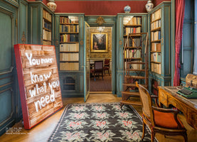 A luxurious, traditional library with red walls and teal-painted, floor-to-ceiling bookshelves. In the foreground, a large, contemporary art piece with a red frame leans against a shelf, displaying the text You never know what you need. The room also features a wooden rolling ladder, a desk with a vintage typewriter, and a floral rug on a hardwood floor. An open doorway reveals an adjoining room with a portrait hanging on the wall.