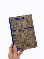 A hand holds a book against a white background. The book cover has a rustic texture made of pressed straw and vibrant blue flower petals. In a bold blue font, the word Unlimited is at the top and the number 023 is at the bottom. The spine of the book is a matching solid blue.