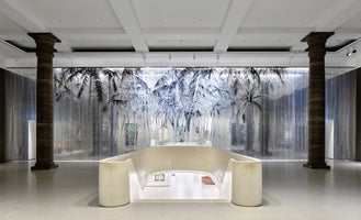 A wide, symmetrical shot of a modern art gallery with a polished floor and white coffered ceiling. The space is framed by two large, dark classical columns. In the center, a curved, cream-colored balustrade surrounds an opening to a lower level. The back wall is covered by a large, backlit, translucent curtain depicting a monochromatic, stylized palm tree forest.