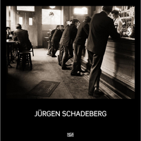 A sepia photograph by Jürgen Schadeberg of men in suits lining a long wooden bar in a pub.