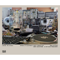 A turntable in the foreground of a music studio packed with a wall of modular synthesizers and a dense tangle of wires.