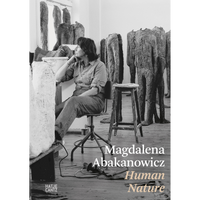 A black and white photograph of the artist Magdalena Abakanowicz sitting in her studio, surrounded by her large, headless figurative sculptures.