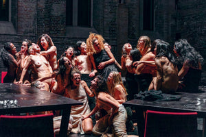 A large group of diverse women, many of whom are topless, are gathered in a dimly lit, brick-walled setting. They are all screaming with expressions of intense anguish and rage, holding onto one another in a powerful, dramatic scene from a theatrical performance.
