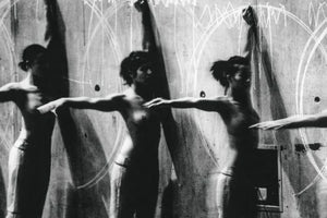A dynamic, grainy black and white photograph capturing a dancer's movements through multiple exposures. The shirtless performer is shown in several overlapping positions with arms outstretched and raised, standing before a rough wall covered with large, sweeping white circles that trace their motion.