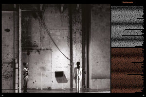 A stark black and white photograph from a book spread showing a performance in a large, raw industrial hall with distressed concrete walls. Two performers stand on opposite sides of the frame. On the left, a woman stands in profile wearing boots and sunglasses. On the right, a woman in briefs stands with her back to the viewer.