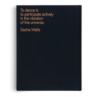 A flat lay photograph of a black, hardbound book on a white background. On the cover, text is printed in a copper-colored sans-serif font. It reads: To dance is to participate actively in the vibration of the universe. Sasha Waltz.