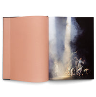 An open book showing a blank, peach-colored page on the left and a full-page photograph on the right. The photo captures a group of contemporary dancers on a dark stage, their expressive poses illuminated by a dramatic, hazy column of light coming from above.