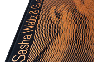 A close-up, angled shot of a black hardcover book with details printed in a shiny, metallic copper color. The spine shows the title Sasha Waltz & Gue, and the front cover features a large portrait of a face rendered in a halftone dot pattern.