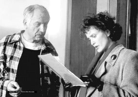 A black-and-white, halftone-style film still from Music Box (1989). A man with a mustache in a plaid shirt and a woman with dark curly hair in a patterned coat stand together, both looking down intently at a document they are holding.