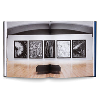 An open art book displays a two-page photograph of a gallery exhibition. Five large, framed black-and-white photographs hang in a row on a white wall above a light wood floor. A black leather L-shaped bench sits in the center of the room, facing the artwork.