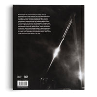 The back cover of an art book, featuring a dramatic, black-and-white, hyperrealistic charcoal drawing by Robert Longo. A brilliant diagonal streak of light cuts through a dark, atmospheric industrial scene. On the left side, there is a block of text describing the exhibition Robert Longo: The Acceleration of History, along with publisher logos, an ISBN, and a barcode at the bottom.