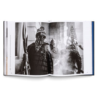 An open book displaying a black-and-white photograph of the January 6th U.S. Capitol insurrection. A person in the foreground wears a beanie and a bandana over their face while holding a flagpole. In the background, amidst smoke, another individual wears a gas mask, and someone else holds up an American flag.