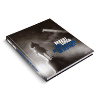 A photograph of the hardcover book Robert Longo: The Acceleration of History, shown at an angle on a white background. The cover features a dramatic, black-and-white image of a silhouetted figure on horseback against a dark, misty sky. The title is printed over the image in large white and bold blue text.