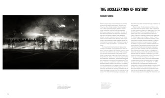 A two-page spread from a book or catalog. On the left is a large, hyperrealistic black and white charcoal drawing by Robert Longo. The artwork shows a nighttime scene with a line of police in riot gear standing in a haze of smoke or tear gas, with bright lights and an explosion in the background. The right page features an essay titled The Acceleration of History by Margaret Andera.
