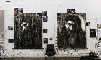 An artist's studio with a white wall smudged with black paint. Two large, heavily textured black and white portraits hang side by side, their subjects' faces partially obscured by vertical streaks. A modern black office chair is centered between the two large pieces, with smaller studies of the portraits tacked to the wall around them.