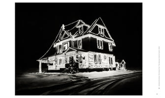 A black and white photograph of a large, multi-story house at night, completely outlined with bright Christmas lights. The house glows against the dark sky, surrounded by snow with tire tracks visible in the foreground.