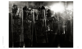 A grainy, black-and-white, close-up photograph of a line of police officers in riot gear at night. They are holding tall, transparent shields that partially obscure their figures and helmets. A bright light flares from the upper right, and the word POLICE is faintly visible through one of the shields.