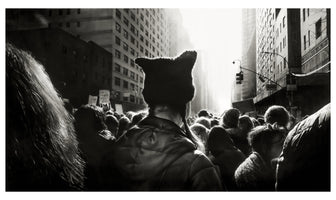 A black and white image from behind a person wearing a cat-eared pussyhat, standing in a large crowd of protesters on a city street. The people are flanked by tall buildings and march toward a bright, hazy light shining down the street, backlighting the scene.