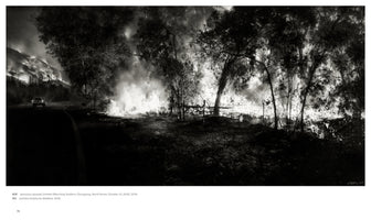 A wide, black and white photograph of a massive wildfire at night. On the left, a single car drives on a dark road with a burning hillside in the distance. To the right, intense flames rage along the roadside, silhouetting trees and filling the air with smoke.