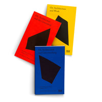 An overhead shot of three books with brightly colored covers, arranged on a white surface. A blue book in the foreground is titled On Architecture and Greenwashing. Behind it are a red book titled On Architecture and the Greenfield and a yellow book titled On Architecture and Work. Each cover features a minimalist design with a large, black, abstract geometric shape.