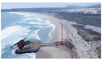 An aerial view of a coastal construction project on a sandy beach with crashing waves. A tall red crane and other machinery are working on a rock jetty, at the end of which a large, red, boxy section of a ship is partially submerged and tilted in the water.