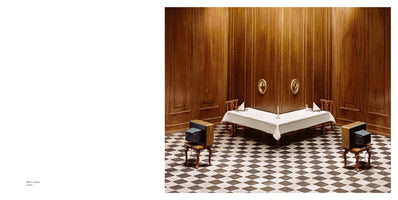 An art photograph of a formal dining room with wood-paneled walls and a black-and-white checkerboard floor. A long table is set for two with a white tablecloth and a single candlestick. In place of people, two vintage television sets sit on small stools, facing each other from opposite sides of the room as if they are the diners.