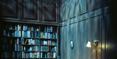 The corner of a dimly lit room with dark wood-paneled walls. On the left, a built-in bookcase is filled with rows of books. On the right, a small, ornate wall sconce with a bell-shaped shade is lit, casting a warm yellow glow on the wall.