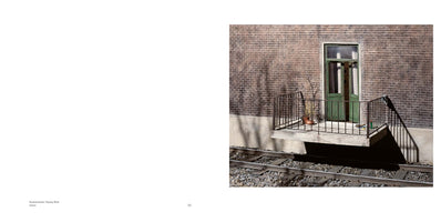 A stark, sunlit photograph of a small concrete balcony with a black railing, jutting out from a brown brick building. A green door leading onto the balcony is slightly ajar. Directly beneath the balcony, a set of train tracks runs along the ground, with strong shadows cast across the scene.