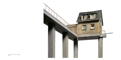 An architectural model of a small, weathered house perched on tall, thick concrete pillars against a white background. A concrete walkway with a rusty railing, also elevated on pillars, leads to the isolated house from the left.