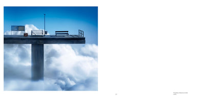 A surreal photograph of a concrete platform resembling a pier or bus stop, supported by a single column and floating high above a sea of white clouds. On the platform, under a clear blue sky, are an empty bench, a lone brown suitcase, a noticeboard, and a lamppost, creating a dreamlike scene of waiting.