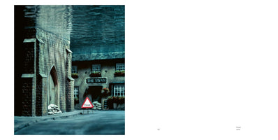A surreal image of a miniature English street scene, with the top half submerged underwater. At street level, a stone building has sandbags piled by its door, and a red triangular sign warns of a flood. In the background, a pub called The Swan is visible below the waterline.