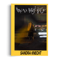 The cover of a book by Sandra Knecht with a bright yellow border. The central image is a nighttime photograph of a person from behind wearing a dark coat and a large, hairy, blonde headdress with two horns. The figure stands on a city street with blurred lights in the background. The title, Home is a Foreign Place, is written in a white, scratchy font at the top.