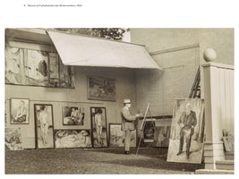 A black and white photograph from 1933 shows the artist Edvard Munch painting in his open-air winter studio. He stands in profile, wearing a suit and hat, working at an easel. He is surrounded by many of his finished paintings, mostly nudes and portraits, which are hung on or leaning against the wall of the building.