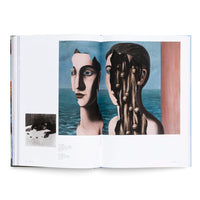 A photograph of an open art book displaying a surrealist painting by René Magritte, The Double Secret, across a two-page spread. The painting shows two female busts against a seascape. The bust on the left is fragmented, with the sky visible through a hole in her face. The bust on the right is broken open, revealing a dark interior with vertical tendrils, each holding a small sphere or bell. A smaller black-and-white photo is on the bottom left page.