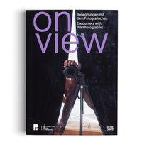 The black cover of a book titled on view in large lavender letters, with the subtitle Encounters with the Photographic. The cover image shows a reflection of a person with dark skin holding a DSLR camera on a tripod, seen in the gap between two dark surfaces. Logos for Pinakothek der Moderne and Hatje Cantz are at the bottom.