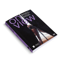 An angled view of a paperback book titled on view, subtitled Encounters with the Photographic, set against a white background. The black cover features a photograph of a person's reflection taking a picture with a camera on a tripod. The title is in large, purple letters, and the book's spine is also purple.