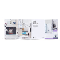 An open exhibition catalog titled on view, Encounters with the Photographic, is spread across a white surface. The pages show a collage of images, including screenshots of computer interfaces, architectural plans, and photographs of people and spaces, all layered over a clean, white interior setting.
