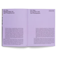 An overhead photograph of an open book showing a two-page spread. The left page has the German title, On View. Begegnungen mit dem Fotografischen. The right page has a lilac header with the English title, On View: Encounters with the Photographic, and authors' names. Both pages feature black text in a two-column layout.