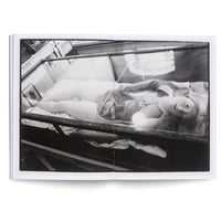 A black and white photograph in an open book showing a female mannequin lying inside a glass display case. The mannequin has blonde hair, a pearl necklace, and looks up towards the viewer while a glowing orb rests on her chest.