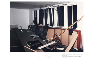A stark, wide photograph of an office interior in complete disarray, created by Thomas Demand. The room is filled with debris, including an overturned large black desk in the foreground and scattered planks of wood. In the background, a row of tall, vertical windows with damaged white frames reveals only blackness. The entire scene is constructed from paper and cardboard, giving it a clean, sterile, and unsettling appearance despite the chaos.