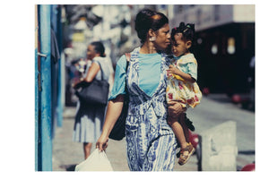 A color street photograph of a Black woman with her hair in an updo, walking on a sunny sidewalk while carrying a young girl on her hip. The woman wears a blue and white patterned jumpsuit and holds a white plastic bag. The child, wearing a floral dress, looks towards the woman's face. The background is slightly blurred, showing another person walking away.