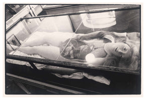 A black-and-white, high-angle photograph of an anatomical model of a woman lying in a glass display case. The lifelike figure has long hair, a pearl necklace, and a doll-like face looking upwards. Her torso is open, exposing a detailed model of her internal organs. The setting appears to be a museum or exhibition.
