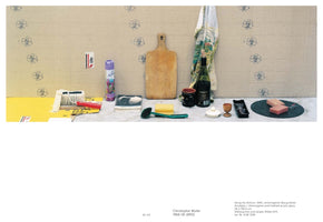 A still-life photograph showing a long row of household items arranged on a shelf against a beige wall patterned with blue recycling symbols. The objects include an air freshener, a wooden cutting board, a bottle of wine, brushes, a sponge, and two pink blocks on a round slate.