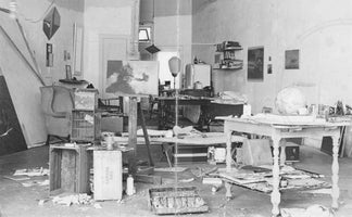 A black and white photograph of a cluttered artist's studio, filled with creative chaos. A painting rests on an easel in the center, surrounded by tables laden with art supplies, a globe, and wooden crates. The floor is strewn with papers, paint, and various objects, indicating an active workspace.