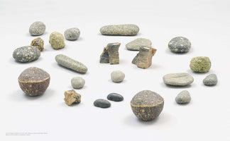 A collection of over twenty stones and sculpted objects arranged on a white surface. The objects vary in shape and texture, including smooth grey pebbles, rough greenish rocks, and angular fragments of layered stone. Two larger, spherical objects are composed of a brown, rough bottom half and a speckled grey top half.