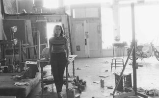 A black and white photograph of a woman in a striped top standing in a spacious and cluttered artist's studio. The room is filled with canvases, an easel, paint supplies, and a scooter is visible in the brightly lit background.