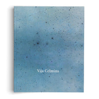 A vertical book cover with the name Vija Celmins in white serif font against a mottled blue background. The background is covered in small black specks, resembling a starry night sky. The book is set against a white surface, casting a soft shadow to its left.